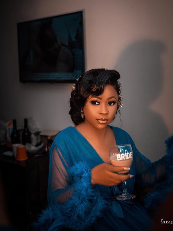 A Woman In A Feathered Blue Robe Holding A Bride Wine Glass In A Dimly Lit Room.